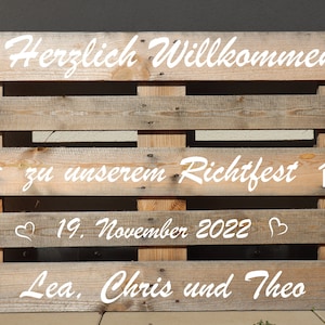 Könnte beinhalten: Ein Holzpaletten-Schild mit weißer Beschriftung, die "Herzlich Willkommen zu unserem Richtfest 19. November 2022 Lea, Chris und Theo" lautet. Das Schild ist mit kleinen Haussymbolen und Herzen verziert.