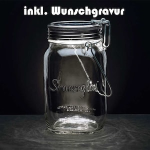 Può includere: Un barattolo di vetro trasparente con un coperchio nero e un manico in metallo. Il barattolo ha le parole "Sonnenglas" stampate sul lato. Il barattolo è illuminato dall'interno e il testo "inkl. Wunschgravur" è stampato sopra il barattolo.