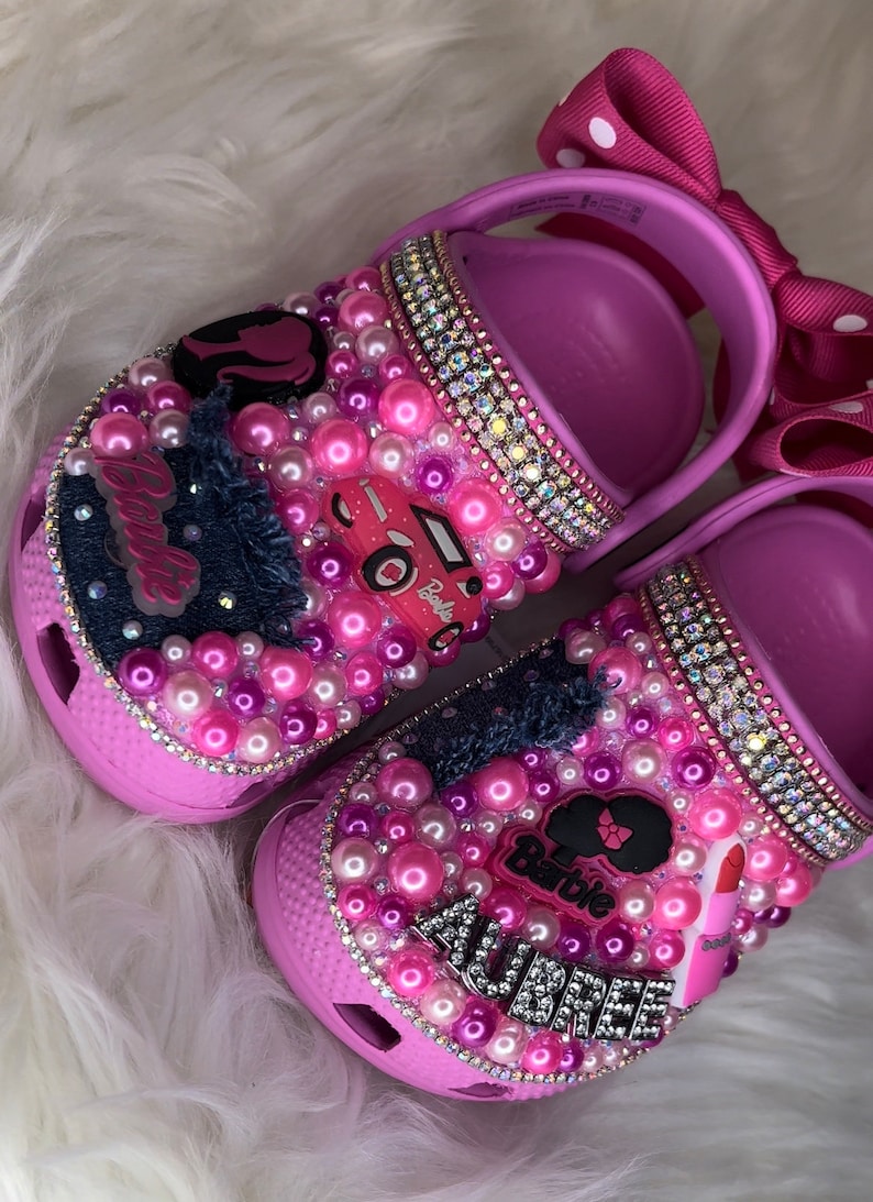 Puede incluir: Zapatos para ni&ntilde;os de color rosa y morado con tem&aacute;tica de Barbie. Los zapatos est&aacute;n decorados con diamantes de imitaci&oacute;n, perlas y un parche de mezclilla. Los zapatos tambi&eacute;n tienen el nombre "Aubree" escrito en ellos.