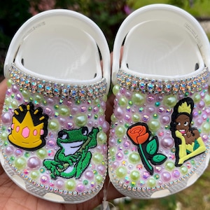 Puede incluir: Un par de zuecos blancos decorados con diamantes de imitaci&oacute;n, perlas y personajes de dibujos animados coloridos. Un zueco presenta una rana, una corona y una hoja verde. El otro zueco presenta una rosa, una princesa y una hoja amarilla.
