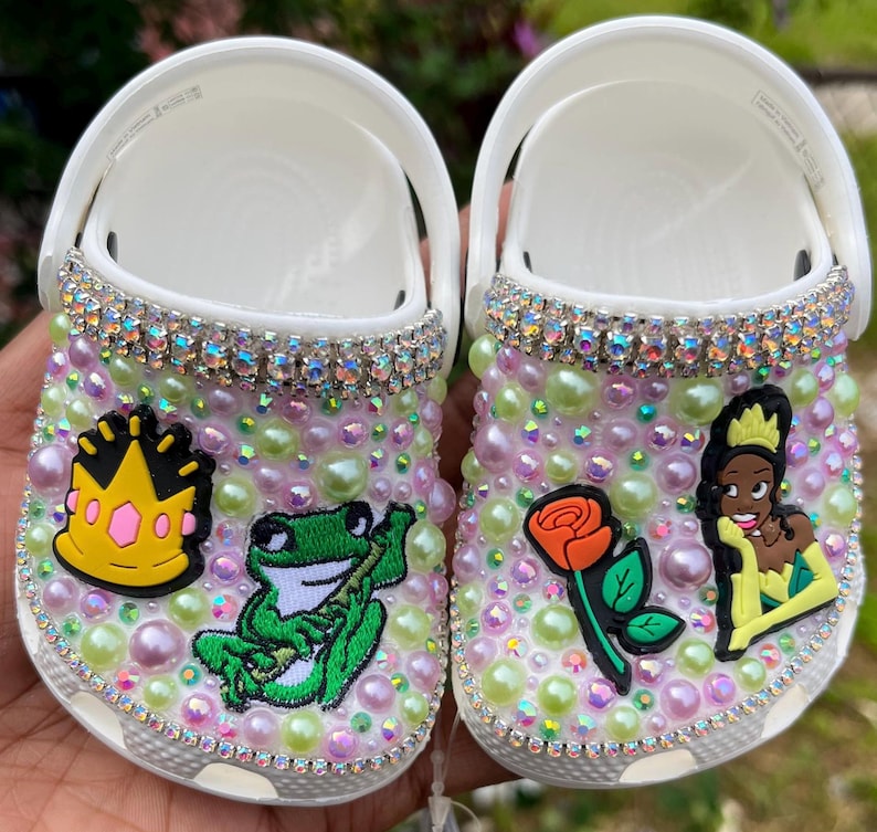 Puede incluir: Un par de zuecos blancos decorados con diamantes de imitaci&oacute;n, perlas y personajes de dibujos animados coloridos. Un zueco presenta una rana, una corona y una hoja verde. El otro zueco presenta una rosa, una princesa y una hoja amarilla.