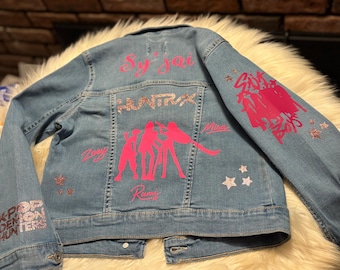 Puede incluir: Chaqueta vaquera azul claro con texto y gráficos rosas y brillantes. La parte trasera presenta las palabras "Sy Jai" y "HUNTX" sobre un gráfico rosa de tres figuras. La manga izquierda dice "K-POP DEMON HUNTERS".