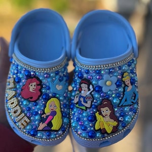 Puede incluir: Zuecos azules personalizados para ni&ntilde;os con un dise&ntilde;o azul y blanco brillante. Los zuecos presentan varios personajes de princesas Disney, incluyendo Ariel, Blancanieves, Bella y Cenicienta. El nombre "Addie" est&aacute; escrito en purpurina dorada en un zueco.