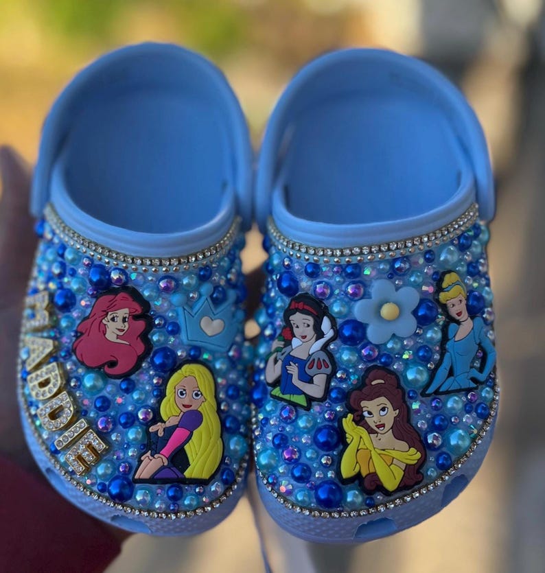 Puede incluir: Zuecos azules personalizados para ni&ntilde;os con un dise&ntilde;o azul y blanco brillante. Los zuecos presentan varios personajes de princesas Disney, incluyendo Ariel, Blancanieves, Bella y Cenicienta. El nombre "Addie" est&aacute; escrito en purpurina dorada en un zueco.