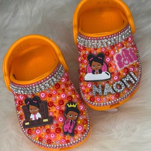 Puede incluir: Zuecos personalizados para ni&ntilde;os de color naranja y rosa con pedrer&iacute;a y un tema de jefa. Los zuecos tienen una imagen de dibujos animados de una ni&ntilde;a con una corona y una ni&ntilde;a con un malet&iacute;n. El nombre "Naomi" est&aacute; escrito en pedrer&iacute;a en un zueco. El texto "The Girl Boss" est&aacute; en el otro zueco.