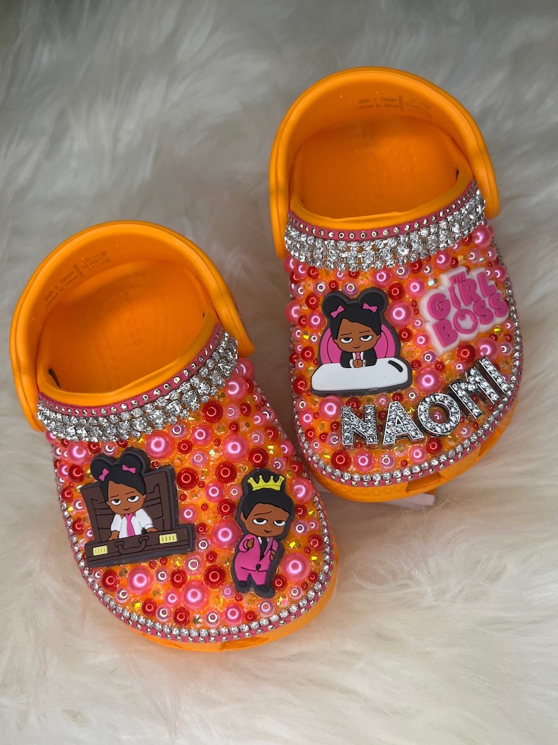 Puede incluir: Zuecos personalizados para ni&ntilde;os de color naranja y rosa con pedrer&iacute;a y un tema de jefa. Los zuecos tienen una imagen de dibujos animados de una ni&ntilde;a con una corona y una ni&ntilde;a con un malet&iacute;n. El nombre "Naomi" est&aacute; escrito en pedrer&iacute;a en un zueco. El texto "The Girl Boss" est&aacute; en el otro zueco.