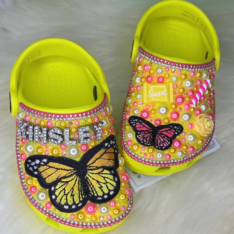 Puede incluir: Un par de zuecos amarillos decorados con pedrer&iacute;a, perlas y aplicaciones de mariposas. Un zueco tiene el nombre "Kinsley" escrito en pedrer&iacute;a y el otro tiene una mariposa rosa y la palabra "Sweet".