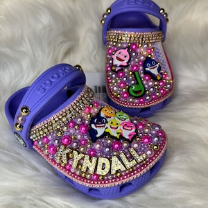 Puede incluir: Zapatos de beb&eacute; de color morado con tem&aacute;tica de tibur&oacute;n beb&eacute;. Los zapatos est&aacute;n decorados con diamantes de imitaci&oacute;n, perlas y una etiqueta de nombre dorada que dice "KYNDALL".