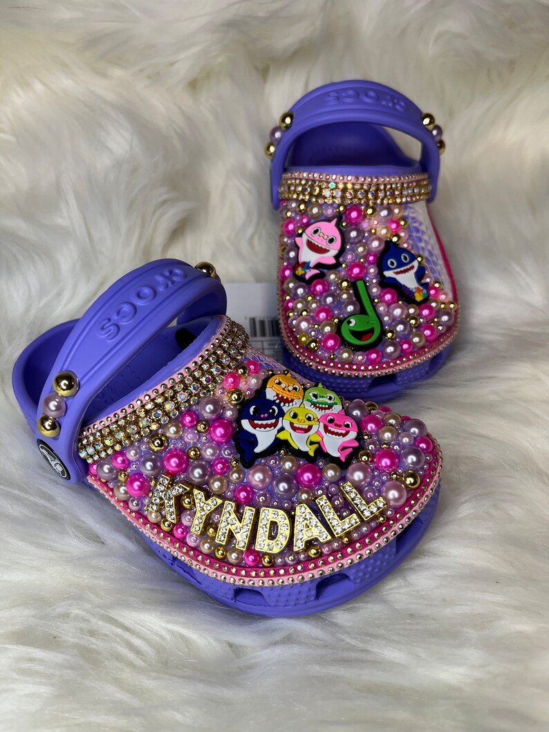 Puede incluir: Zapatos de beb&eacute; de color morado con tem&aacute;tica de tibur&oacute;n beb&eacute;. Los zapatos est&aacute;n decorados con diamantes de imitaci&oacute;n, perlas y una etiqueta de nombre dorada que dice "KYNDALL".