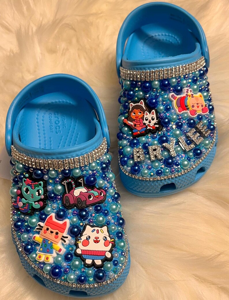 Puede incluir: Crocs azules decoradas con diamantes de imitaci&oacute;n y abalorios de personajes de dibujos animados de colores. El nombre "BRYLER" est&aacute; escrito en diamantes de imitaci&oacute;n en el zapato derecho.