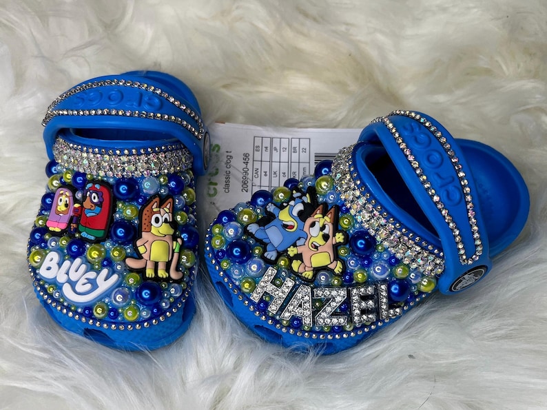 Puede incluir: Un par de zuecos azules para ni&ntilde;os decorados con diamantes de imitaci&oacute;n y personajes de dibujos animados. Los zuecos est&aacute;n personalizados con los nombres "Bluey" y "Hazel".