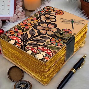 May include: A decorative journal with a floral and mushroom design. The cover features colorful flowers, mushrooms, and a dragonfly. The pages have a golden edge, and the journal is secured with a metal clasp. A black pen rests beside the journal.