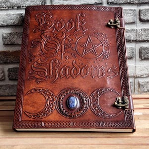 Puede incluir: Un diario de cuero marrón con las palabras "Book of Shadows" grabadas en relieve en la portada, junto con un pentagrama y diseños de luna creciente. El diario tiene un cierre de broche y una piedra azul en el centro.