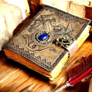 May include: A detailed, antique-style journal with a dragon design on the cover. The cover is tan with a blue gemstone and a metal clasp. A red feather quill rests nearby, suggesting a fantasy or historical theme.