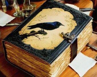 Può includere: Un grande libro in stile antico con copertina nera e illustrazione di un corvo. Il libro ha una chiusura a fibbia e pagine ingiallite. Il corvo è appollaiato su un ramo, su uno sfondo simile a pergamena. Il libro è su un tavolo di legno con altri oggetti vintage.