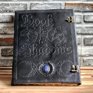 May include: A black leather "Book of Shadows" with embossed lettering and symbols, including a pentagram and crescent moons. It has a central blue stone and metal clasps. The book is displayed against a brick wall.
