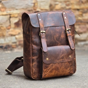 May include: A brown leather backpack with a distressed finish. The bag features a flap closure, adjustable straps, and a front pocket with a snap closure. The backpack has brass-colored hardware and a single shoulder strap.