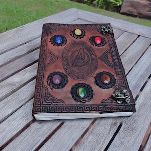 Puede incluir: Un diario de cuero marrón con un logotipo de los Vengadores en relieve y un borde decorativo. La portada presenta seis gemas de colores incrustadas. El diario está abierto, revelando páginas blancas, y descansa sobre una mesa de madera.