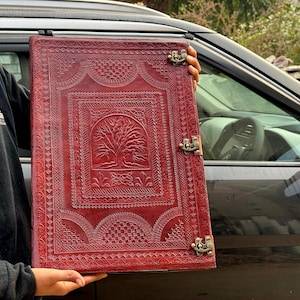 Può includere: Un grande diario rettangolare in pelle rossa con intricati disegni in rilievo. La copertina presenta un motivo centrale ad albero e bordi decorativi. Il diario è assicurato con due fermagli in metallo in stile antico.