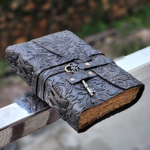 May include: A dark brown leather journal with an embossed floral pattern. The journal is secured with a strap and a decorative key closure. The aged pages are visible, suggesting a vintage or antique style.