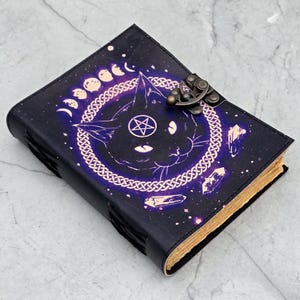May include: A dark blue leather journal with a cat design, a pentagram, and moon phases. The cover features a metal clasp and a Celtic knot border around the cat's face. The pages have a vintage, aged appearance.