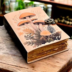 Puede incluir: Un diario encuadernado en cuero con una ilustración detallada de setas y follaje de color naranja. La cubierta tiene un cierre de metal. Las páginas tienen un aspecto vintage. El diario está sobre una superficie de madera.