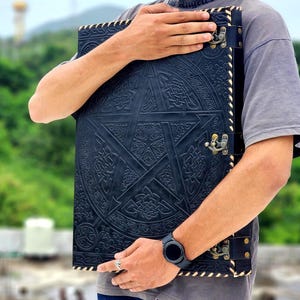 May include: A large, black leather journal with a raised pentagram design and intricate Celtic knotwork. The journal has brass-colored clasps and a stitched border. The cover is dark blue and the pages are not visible.