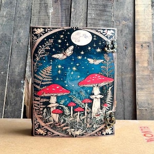 May include: A decorative journal with a fantasy-themed cover. The cover features a night sky with a full moon, butterflies, and red mushrooms. The journal has a dark border and a metal clasp.