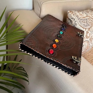 Puede incluir: Un diario de cuero marrón con un diseño en relieve y una fila de piedras preciosas de colores. El diario tiene un interior negro, costuras blancas y cierres metálicos. La cubierta presenta siete piedras preciosas.