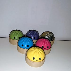 May include: A collection of six small, round, colourful figurines shaped like steamed buns, each resting in a tan circular base. The buns are green, purple, silver, blue, pink, and yellow, all featuring black painted eyes and a small smiling mouth.