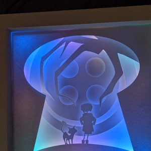 May include: A backlit art piece featuring a silhouette of a girl, a cat, and a large, looming figure within a keyhole shape. The artwork uses layered paper to create depth, with blue, purple, and green lighting.