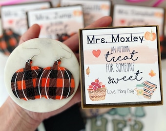 May include: A pair of pumpkin-shaped earrings with a black and orange plaid pattern. The earrings are displayed on a white surface next to a gift box with text that reads "An autumn treat for someone sweet."