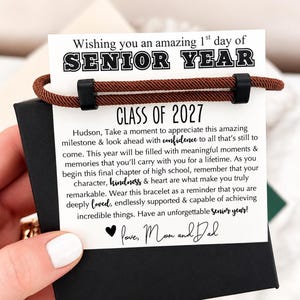 May include: A brown cord bracelet with two black sliding beads attached to a white card. The card features the text: Wishing you an amazing 1st day of SENIOR YEAR, CLASS OF 2027, and a message for Hudson, signed with a heart and Love, Mom and Dad.