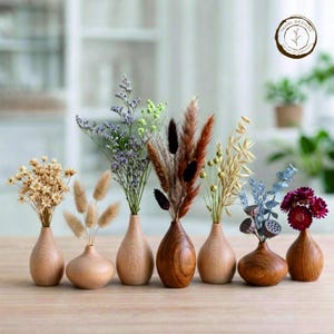 Op de afbeelding: Een verzameling van zeven kleine houten vazen, elk met gedroogde bloemen en grassen. De vazen hebben een natuurlijke afwerking en variëren in vorm en houtnerf. De bloemen omvatten beige, paarse en bruine variëteiten, gerangschikt voor een decoratieve weergave.