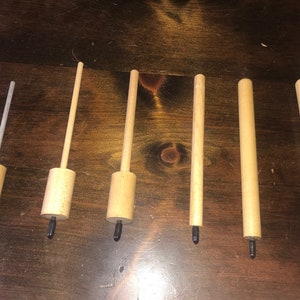 May include: A set of six wooden tools arranged in a row, each featuring a cylindrical wooden handle and a protruding black tip at the base. Three of the tools have thin, extended wooden shafts, one has a thin clear rod, and two are solid cylinders.