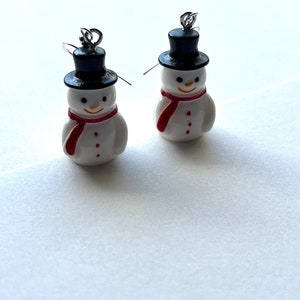 May include: A pair of snowman earrings with black hats, red scarves, and white bodies. The earrings are made of polymer clay and have silver hooks.