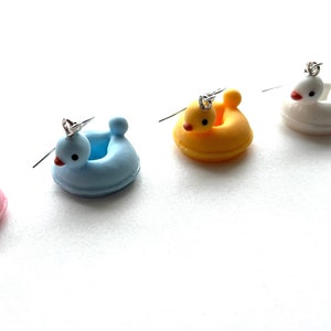 May include: Five pairs of colorful duck-shaped earrings. Each earring features a duck with a crown on its head, sitting in a small inflatable ring. The ducks are pink, blue, yellow, white, and purple.