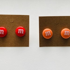 K&ouml;nnte beinhalten: Zwei Paar Ohrstecker. Ein Paar ist rot mit einem wei&szlig;en "M" auf jedem Ohrstecker. Das andere Paar ist pfirsichfarben mit einem wei&szlig;en "M" auf jedem Ohrstecker.