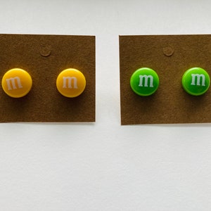 K&ouml;nnte beinhalten: Zwei Paar Ohrringe, ein Paar ist gelb mit einem wei&szlig;en "M" auf jedem Ohrring, das andere Paar ist gr&uuml;n mit einem wei&szlig;en "M" auf jedem Ohrring.