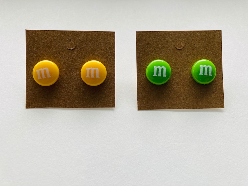 K&ouml;nnte beinhalten: Zwei Paar Ohrringe, ein Paar ist gelb mit einem wei&szlig;en "M" auf jedem Ohrring, das andere Paar ist gr&uuml;n mit einem wei&szlig;en "M" auf jedem Ohrring.
