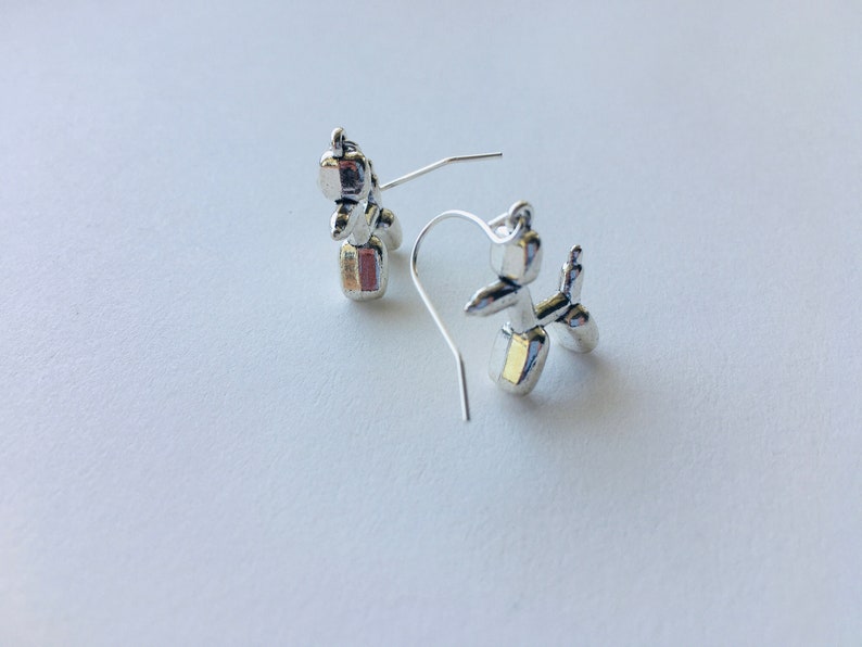 May include: A pair of silver balloon animal earrings. The earrings are shaped like a dog and a cat. The dog is silver with a gold accent. The cat is silver with a pink accent.