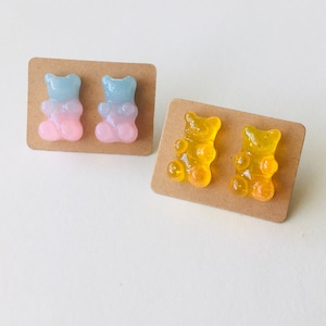 May include: Two pairs of gummy bear earrings. One pair is blue and pink, the other is yellow. The earrings are on brown card stock.
