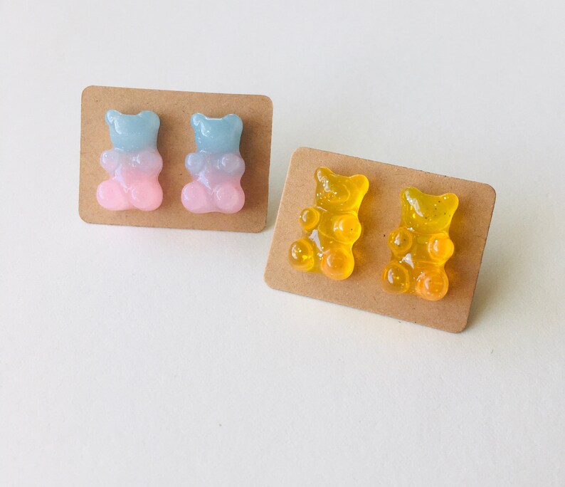 May include: Two pairs of gummy bear earrings. One pair is blue and pink, the other is yellow. The earrings are on brown card stock.