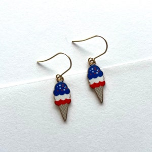 May include: A pair of gold dangle earrings featuring a red, white, and blue ice cream cone design with sprinkles.