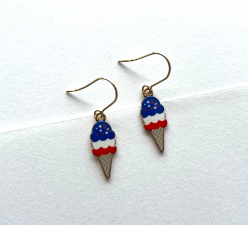 May include: A pair of gold dangle earrings featuring a red, white, and blue ice cream cone design with sprinkles.
