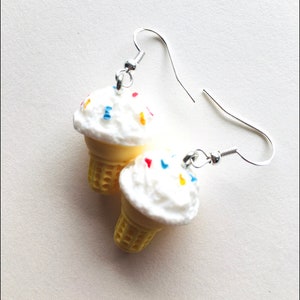 May include: A pair of silver dangle earrings featuring a white ice cream cone with colourful sprinkles. The ice cream cone is made of plastic and is a fun and whimsical accessory.