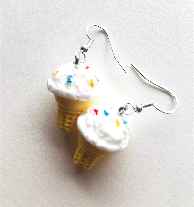 May include: A pair of silver dangle earrings featuring a white ice cream cone with colourful sprinkles. The ice cream cone is made of plastic and is a fun and whimsical accessory.