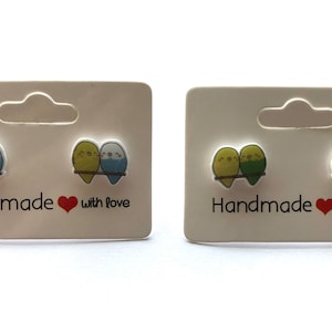 May include: Two pairs of handmade earrings featuring a pair of cartoon parakeets on each earring. The parakeets are green and yellow on one pair and blue and yellow on the other pair. The earrings are on a white card with the text "Handmade with love".