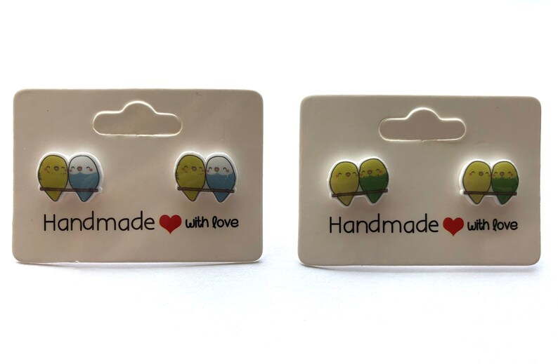 May include: Two pairs of handmade earrings featuring a pair of cartoon parakeets on each earring. The parakeets are green and yellow on one pair and blue and yellow on the other pair. The earrings are on a white card with the text "Handmade with love".