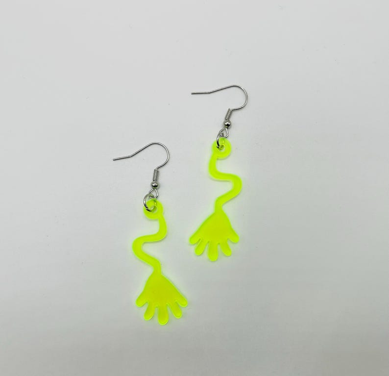 May include: A pair of neon yellow dangle earrings with a hand-shaped design at the bottom. The earrings have a silver hook.